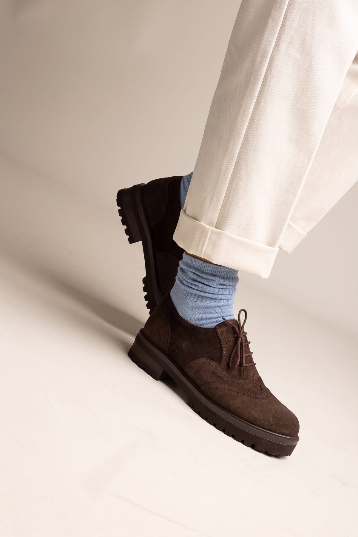 Model mid-step in brown suede brogue shoes with laces and thick lug sole seen from the side with light blue socks and cream trousers