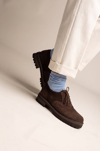 Model mid-step in brown suede brogue shoes with laces and thick lug sole seen from the side with light blue socks and cream trousers