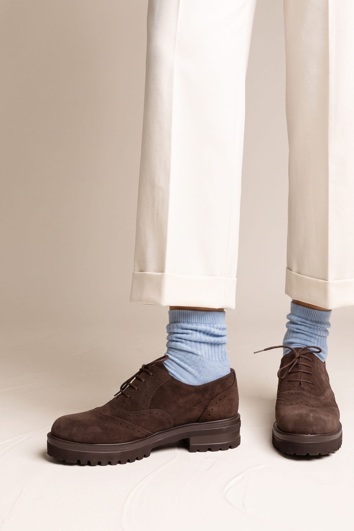 Model wearing brown suede brogue shoes with chunky lug sole and lace detail one foot forward light blue socks and cream trousers