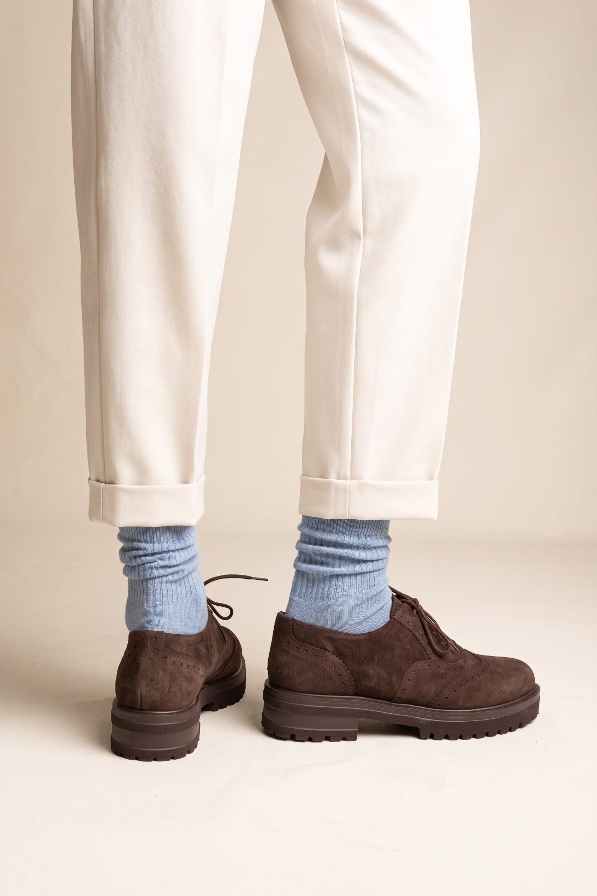 Model wearing brown suede lace up brogue shoes with chunky sole and light blue socks under cream cuffed trousers feet seen from the front