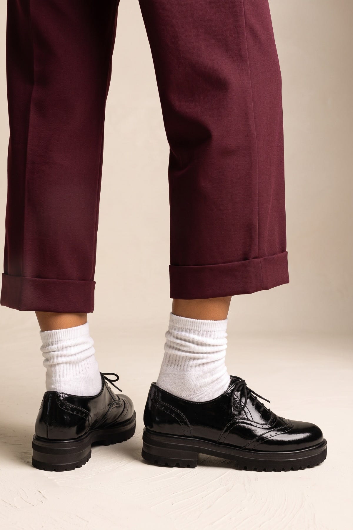 A person wearing dark burgundy cropped trousers, white crew socks, and black leather lace-up shoes with chunky black soles, standing against a plain beige studio background.