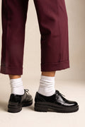 A person wearing dark burgundy cropped trousers, white crew socks, and black leather lace-up shoes with chunky black soles, standing against a plain beige studio background.