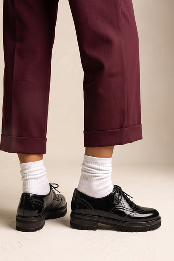 A person wearing dark burgundy cropped trousers, white crew socks, and black leather lace-up shoes with chunky black soles, standing against a plain beige studio background.