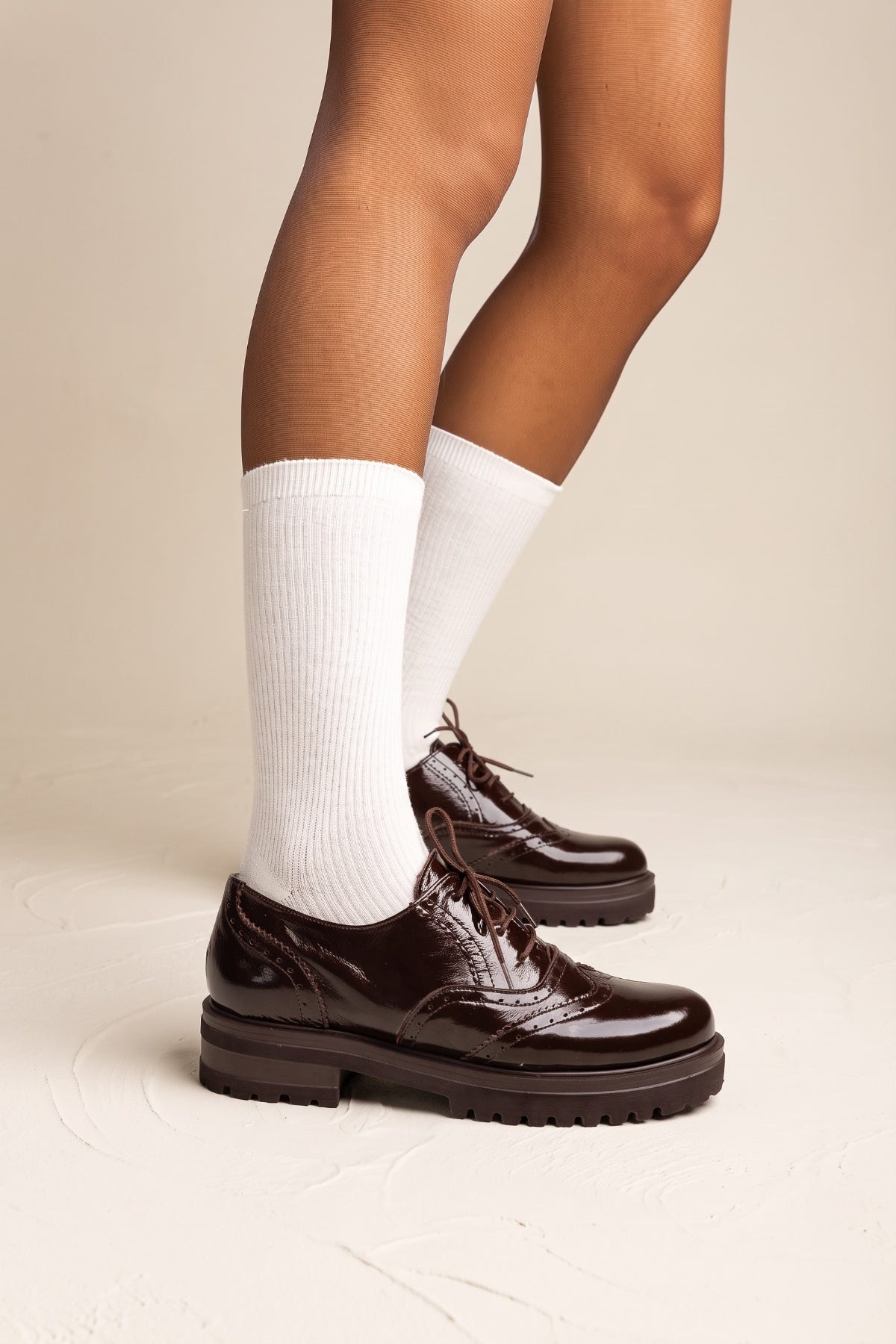 model wearing glossy dark brown lace up brogues with chunky soles and white socks, seen from the side