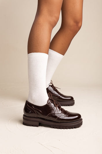 model wearing glossy dark brown lace up brogues with chunky soles and white socks, seen from the side