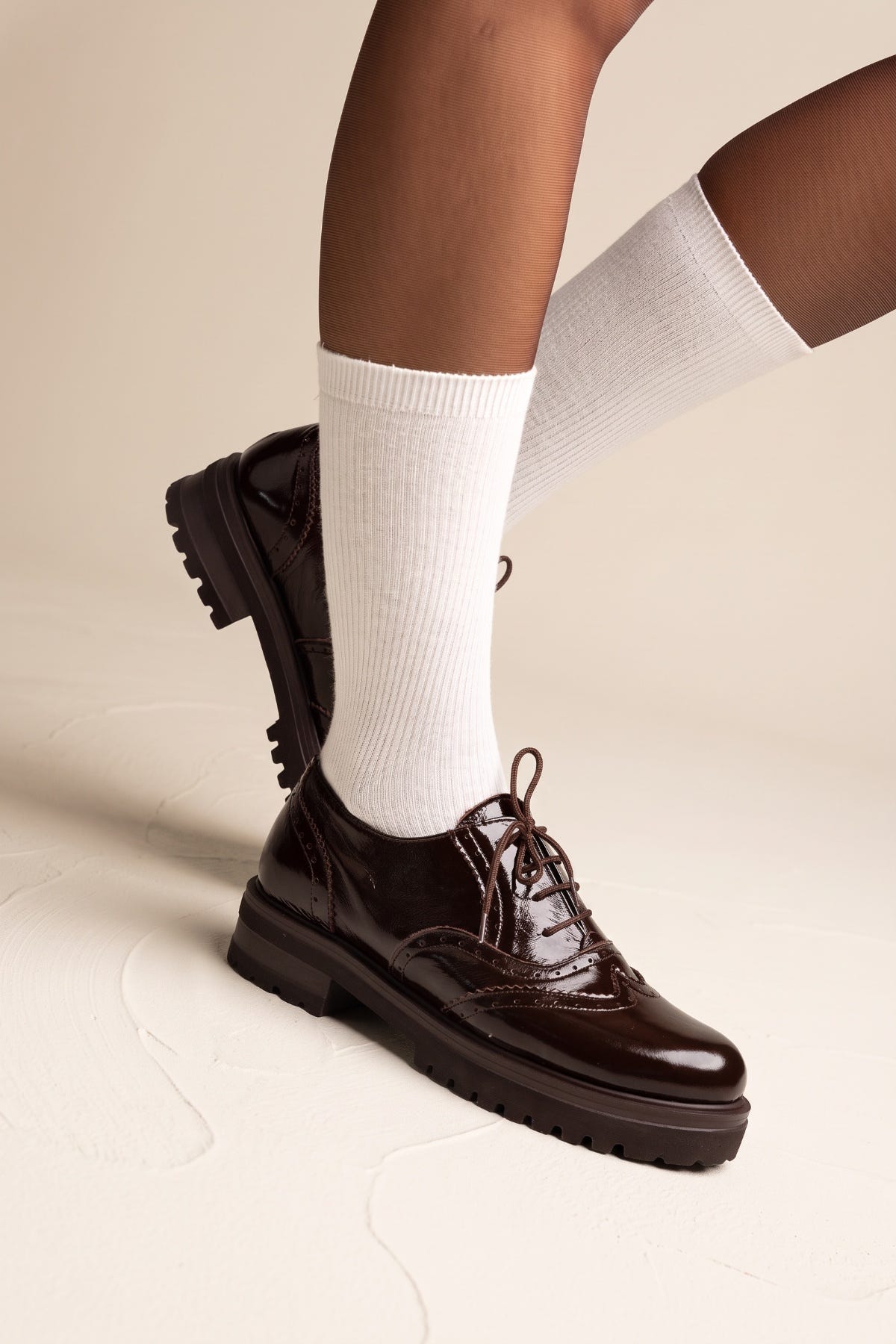 model wearing glossy dark brown lace up brogues with white ribbed socks, one foot lifted to show the sole
