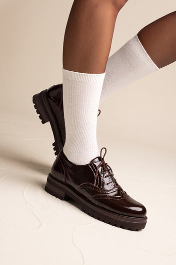 model wearing glossy dark brown lace up brogues with white ribbed socks, one foot lifted to show the sole