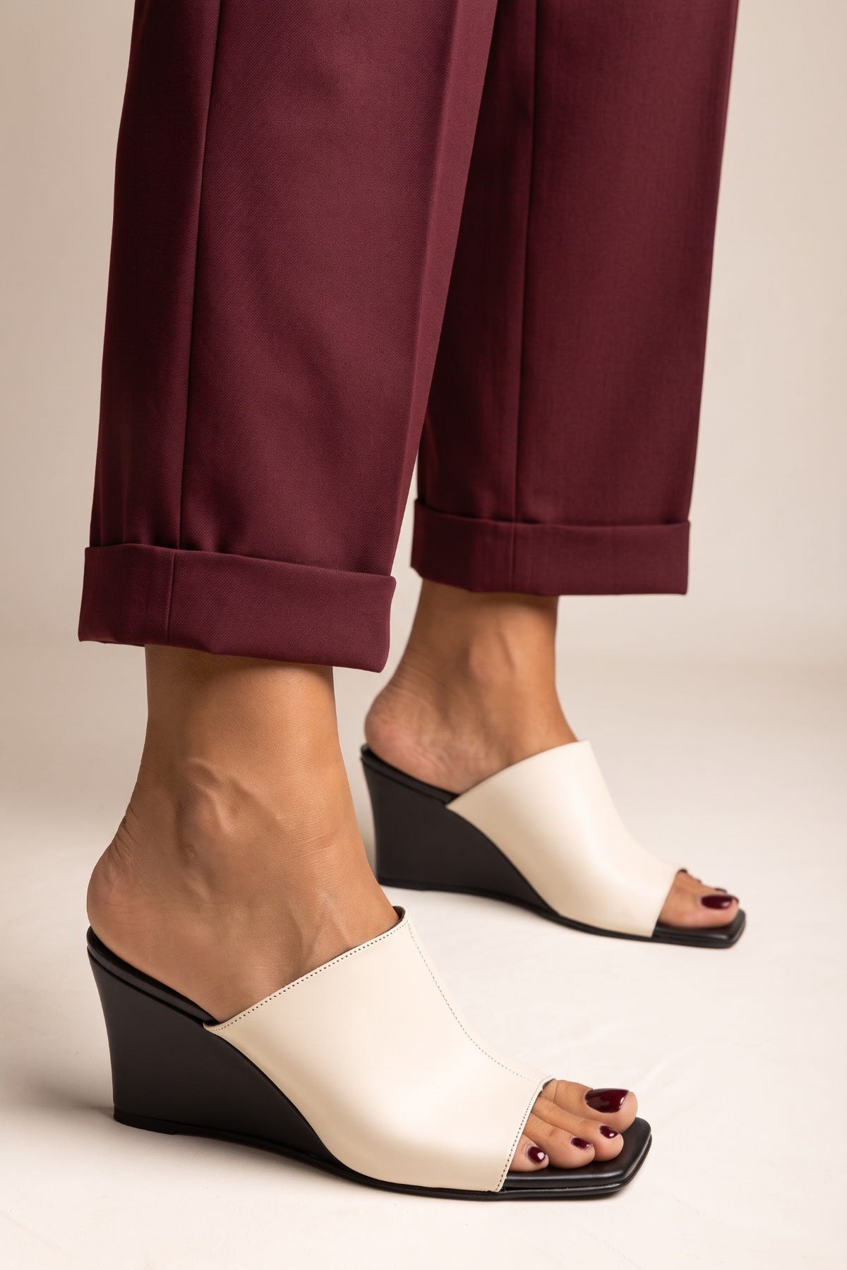 Model feet in beige leather wedge heel mules with black heel and square toes beside burgundy trousers