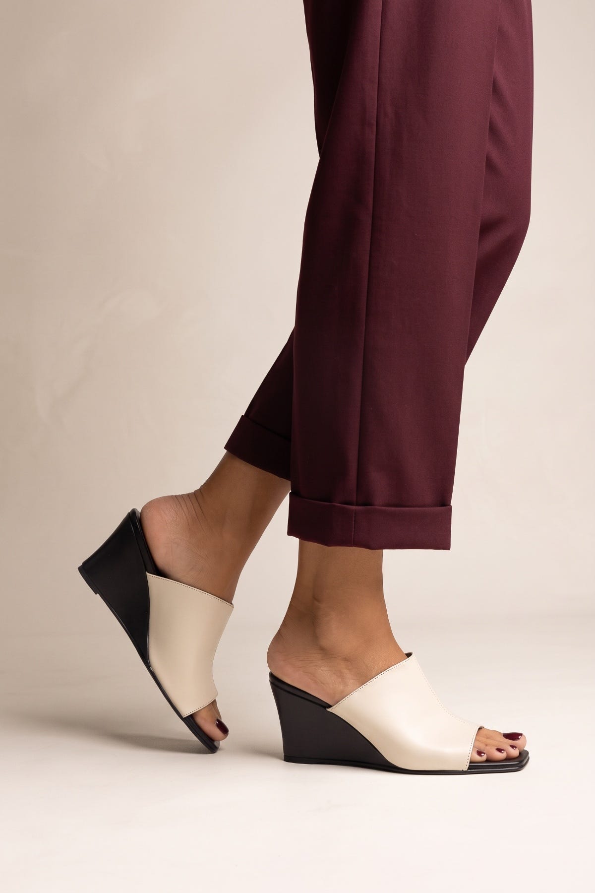 Model wearing beige and black wedge heel mules with square toes and burgundy trousers