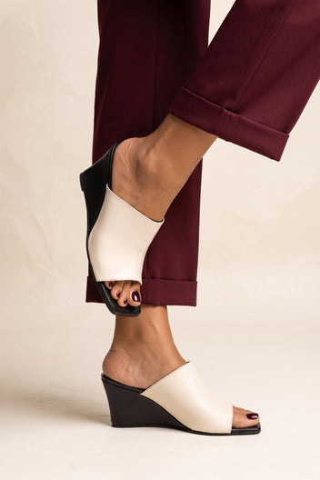 Side angle of model feet in beige and black wedge mules with square toes and burgundy trousers