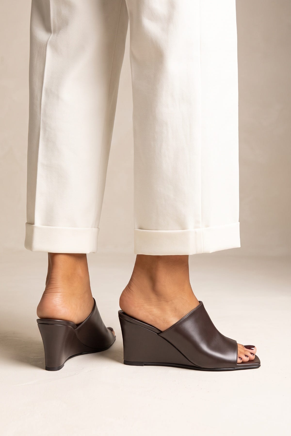 Model wearing brown matte wedge heel mules with square toes and cream cuffed trousers