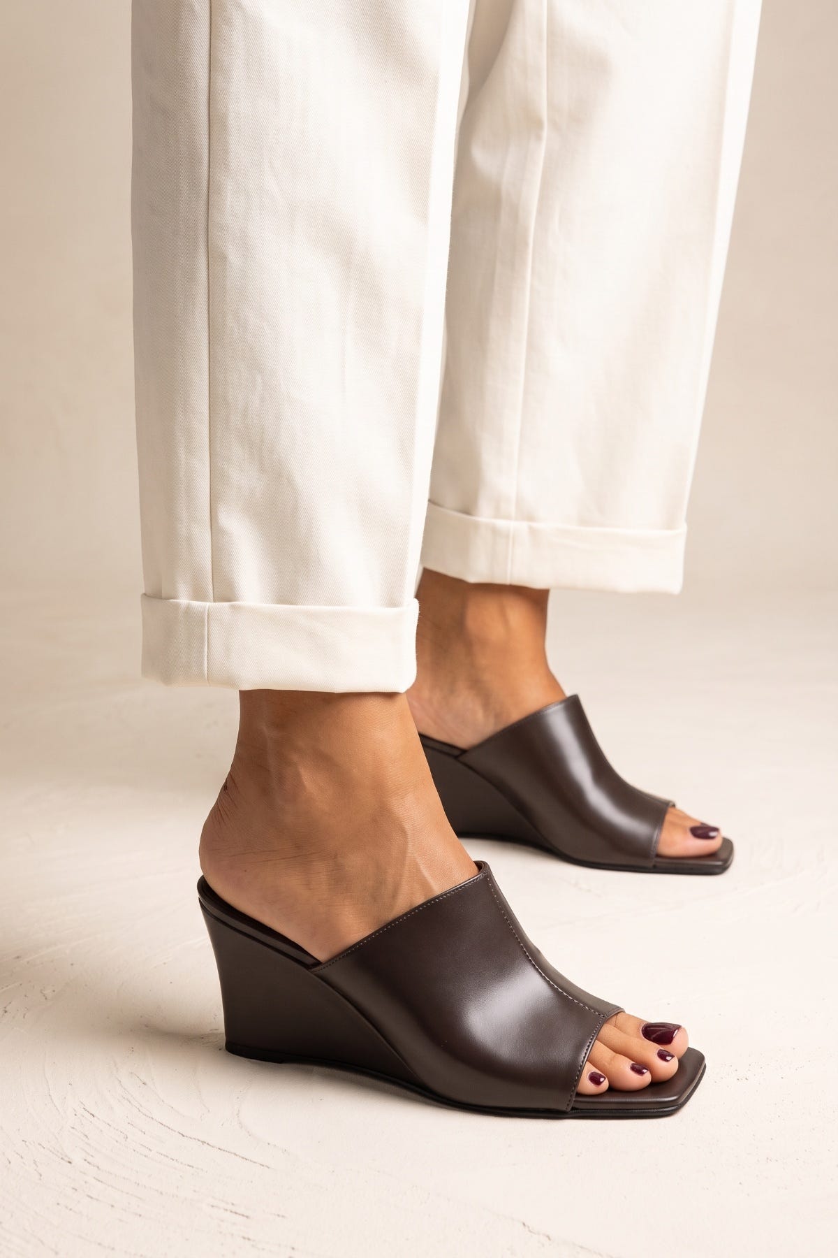 Model in cream rolled cuff trousers wearing brown matte leather wedge mules with an open square toe, front-facing stance