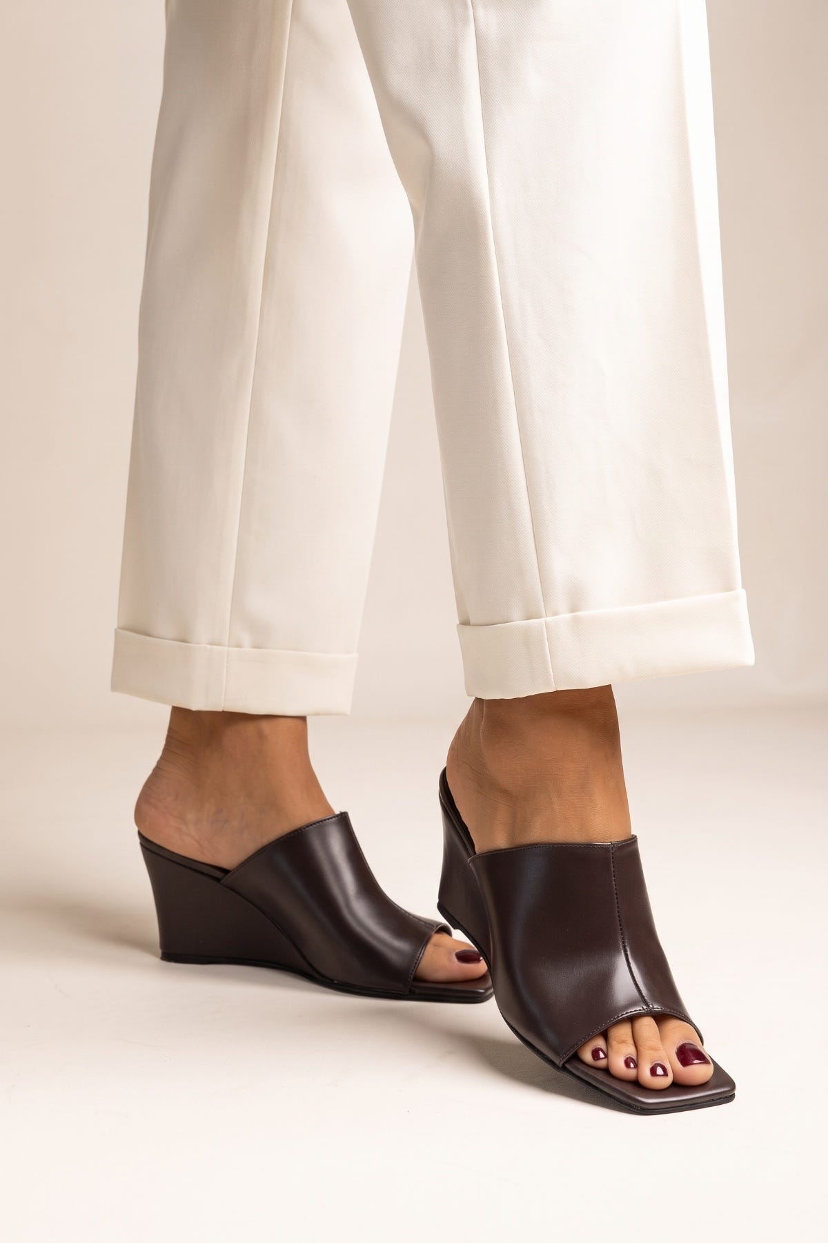 Model wearing brown matte square toe wedge mules with open toes and cream cuffed trousers from the front