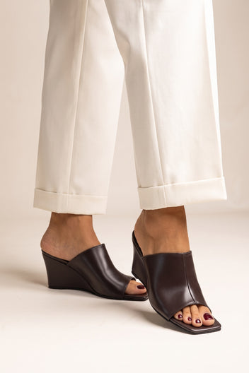 Model wearing brown matte square toe wedge mules with open toes and cream cuffed trousers from the front