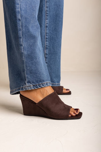Model wearing brown suede square toe wedge heel mules with a low heel and wide leg blue jeans