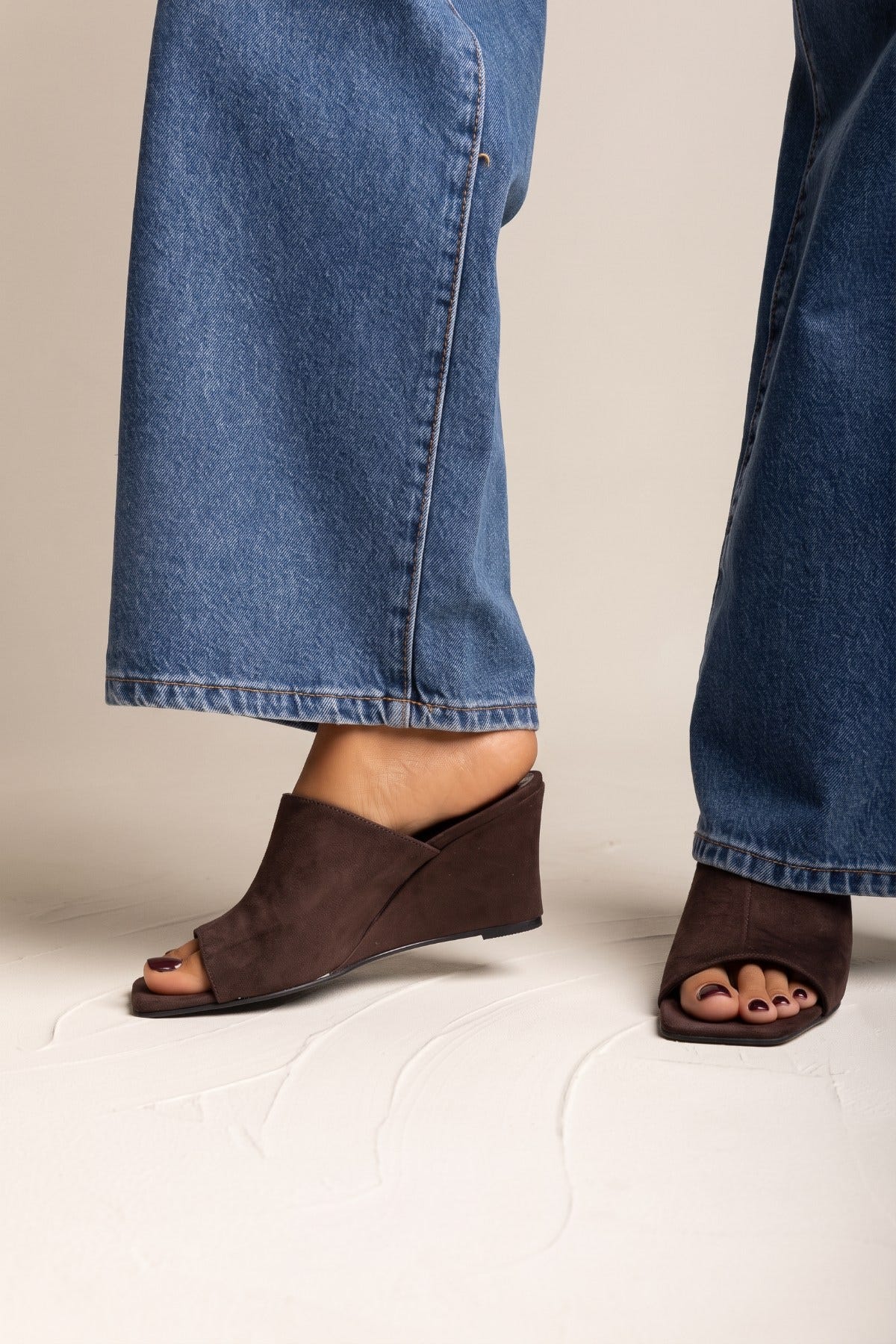 Brown suede wedge heel mules with open square toe on model feet beside wide blue jeans