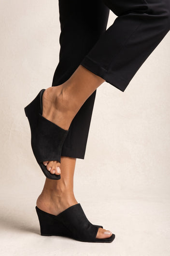 Side view on model of black suede wedge mules with open square toe and angled wide band, solid wedge heel
