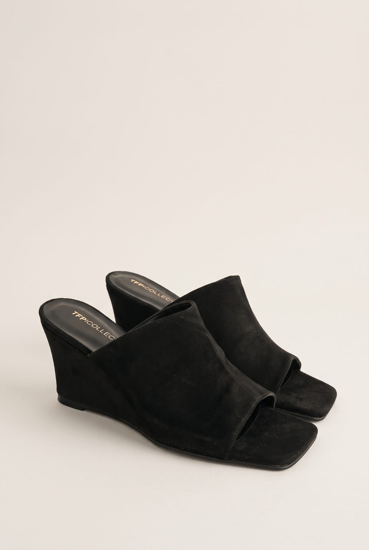 Three-quarter view of a pair of black suede wedge mules with square open toes and wide vamp bands