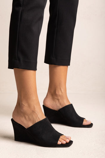 Front view on model of black suede wedge mules with wide diagonal strap, open square toe and high wedge heel
