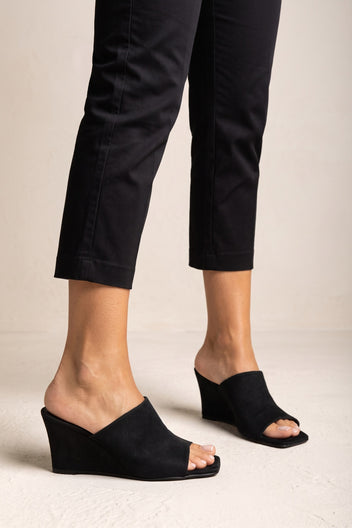 Black suede wedge mule with an asymmetric wide band, open square toe and a solid wedge heel, worn on model