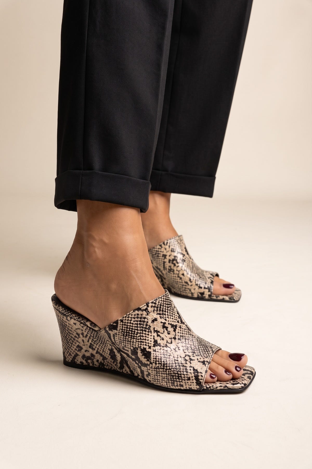 Beige and brown snake print wedge mules with a square toe worn by model in black cuffed trousers, front view of feet