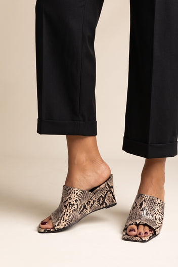 Model wearing beige snake print square toe wedge mules with black cuffed trousers, feet apart, three-quarter front view
