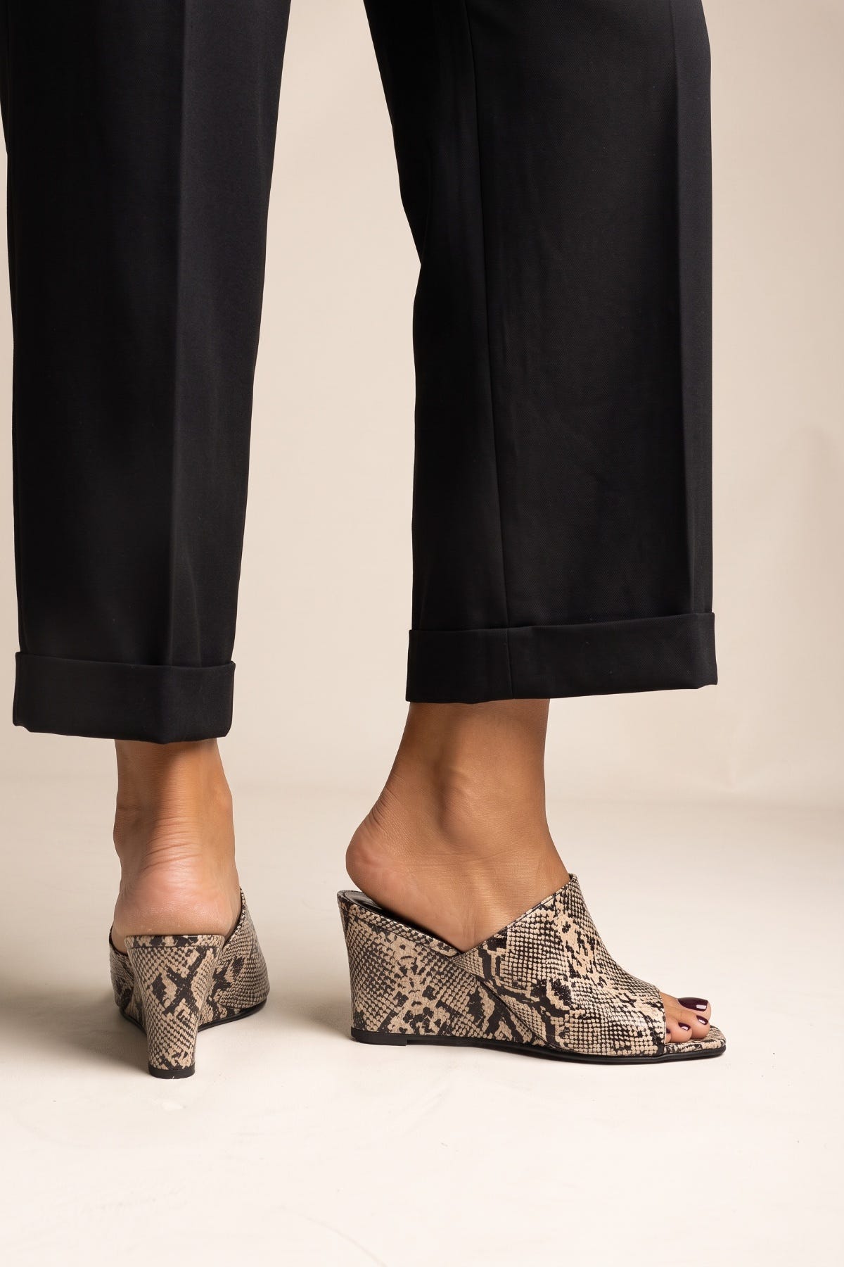 Model in black cuffed trousers standing in beige snake print wedge mules with square toes, full view of both feet