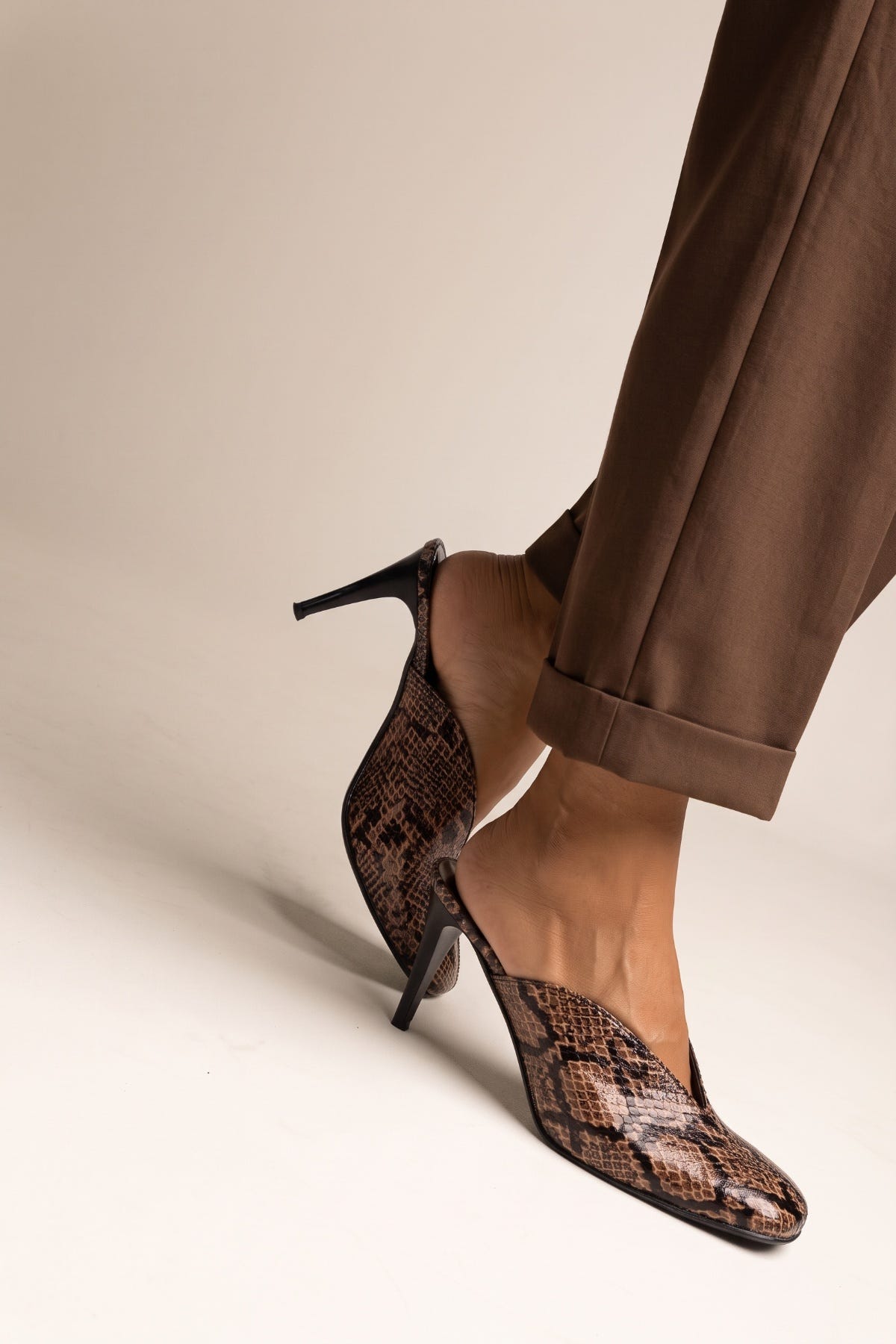 model wearing brown snake print stiletto mules with cuffed trousers, one foot raised to show heel