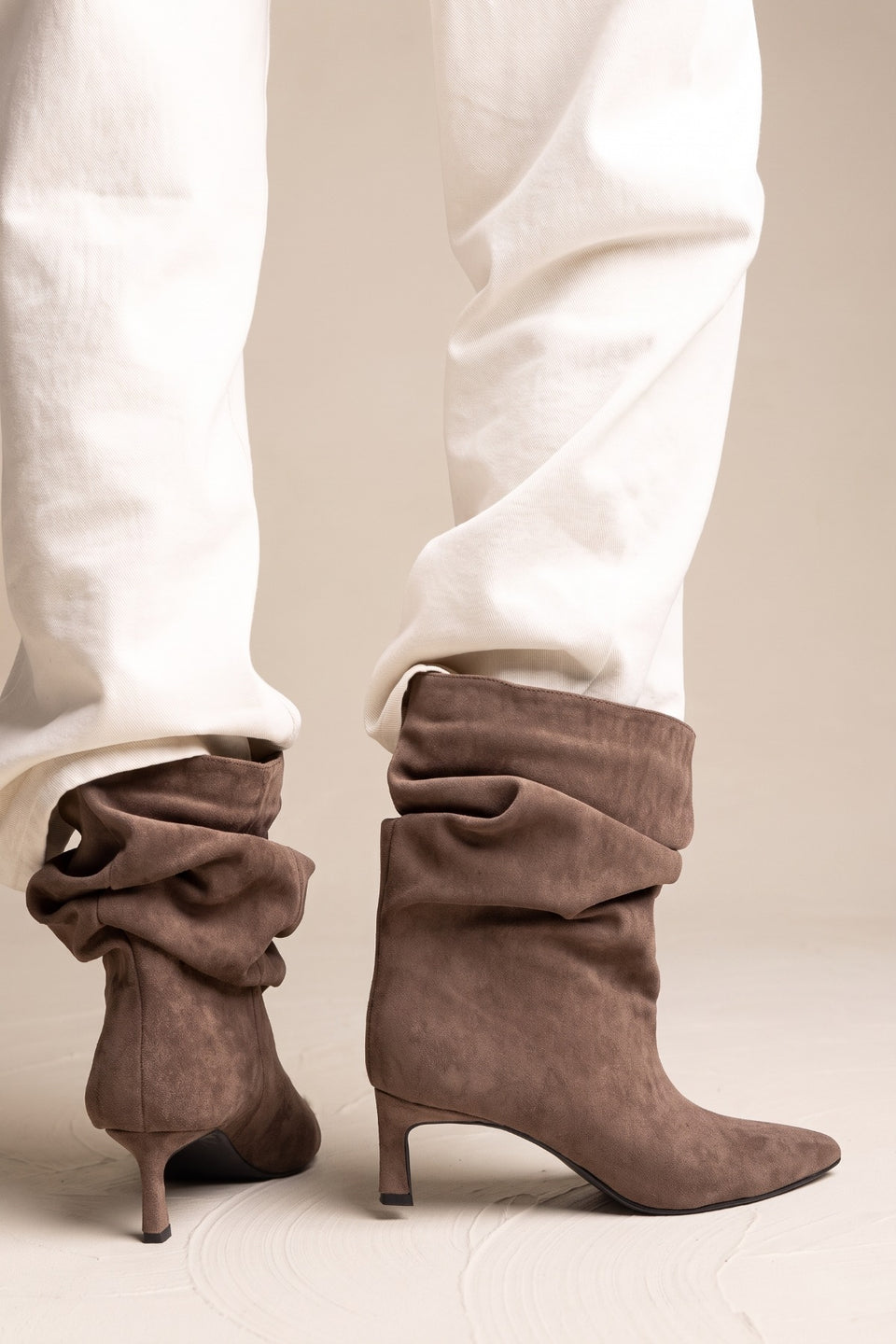 Model stands in white trousers with taupe suede ruched kitten heel boots shown from a front view