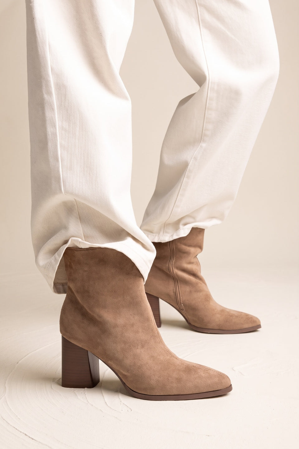 Model in white trousers wearing taupe suede pointed toe ankle boots with block heels