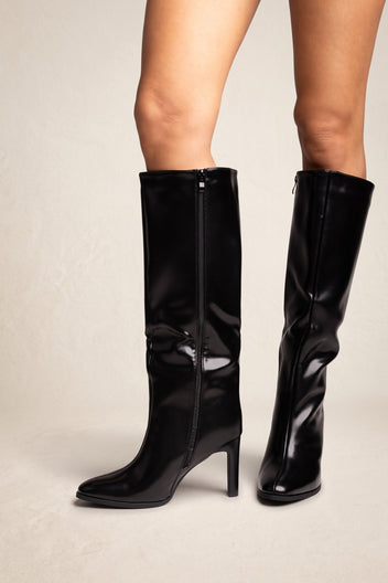 Model wearing black knee-high boots standing with feet apart, showing the side zipper on the inner shaft and pointed toes with block heels