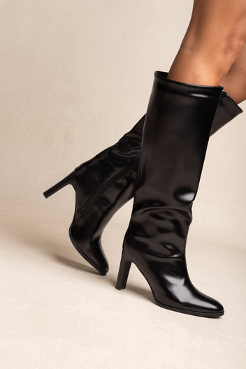 Model wearing black leather-look knee-high boots with one leg lifted and bent, showing the pointed toe and tapered block heel from the side
