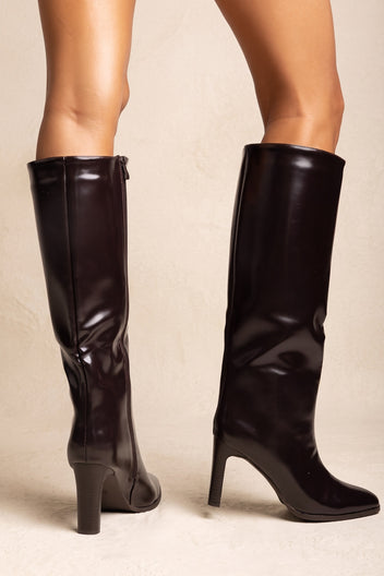 Model wearing dark brown knee-high boots with block heels and back zips seen from behind against a soft beige wall