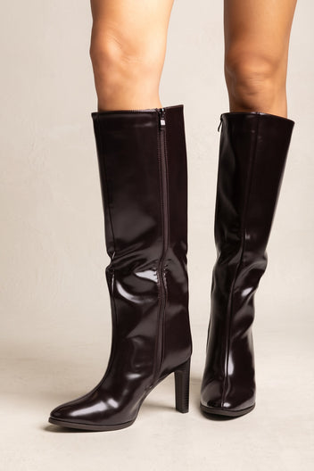 Model wearing dark brown knee-high boots with block heels and side zips seen from the front on a beige floor