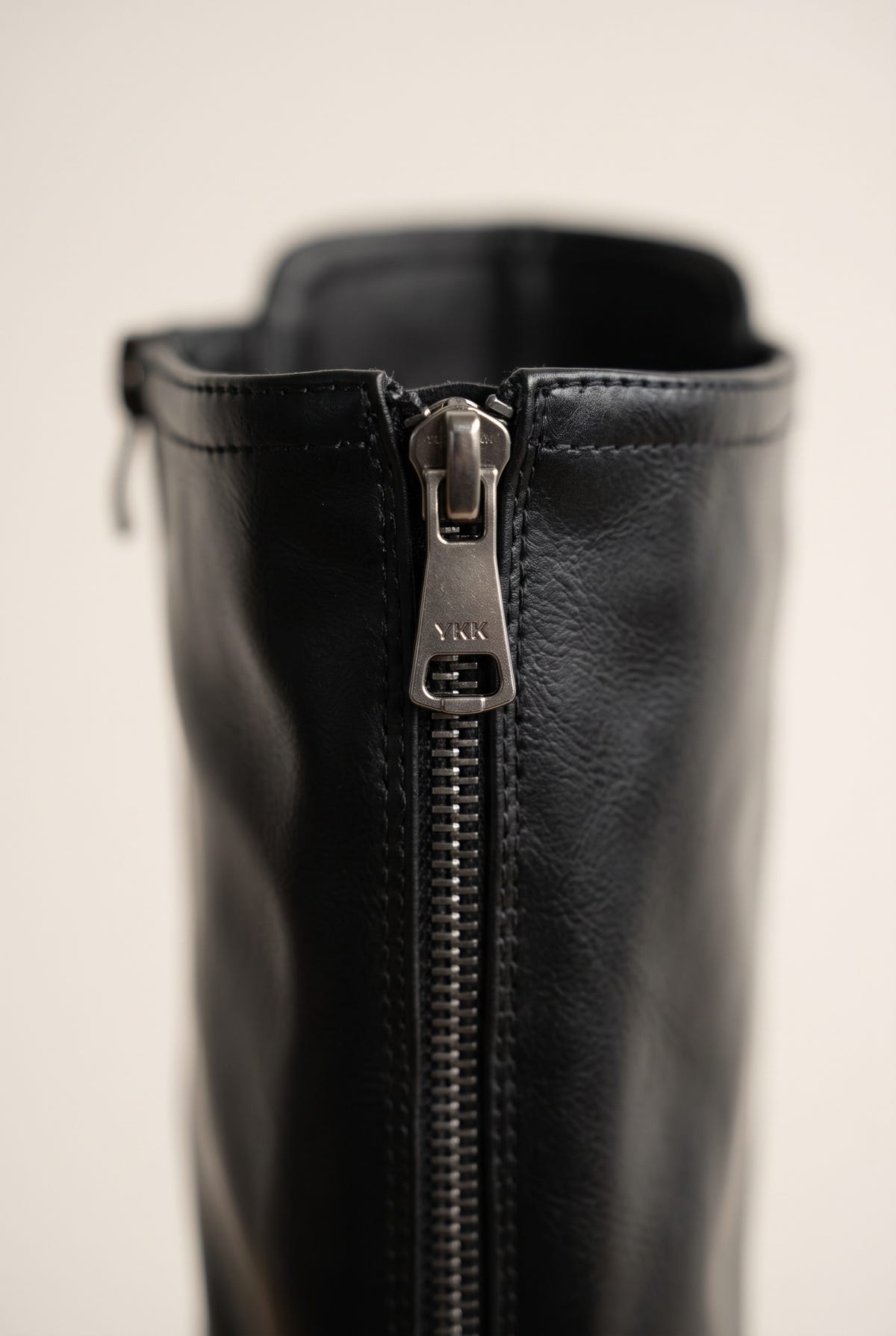 Close-up of a metal zipper pull on the black faux leather shaft of a knee-high boot with a smooth textured finish