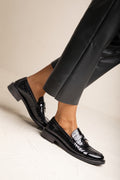 Glossy black croc-embossed loafer with metal bar detail, side view on model in black leather pants