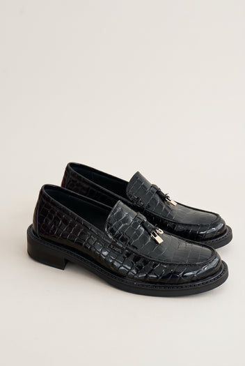 Pair of black croc-embossed loafers in three-quarter view, metal charm on the strap and low chunky soles