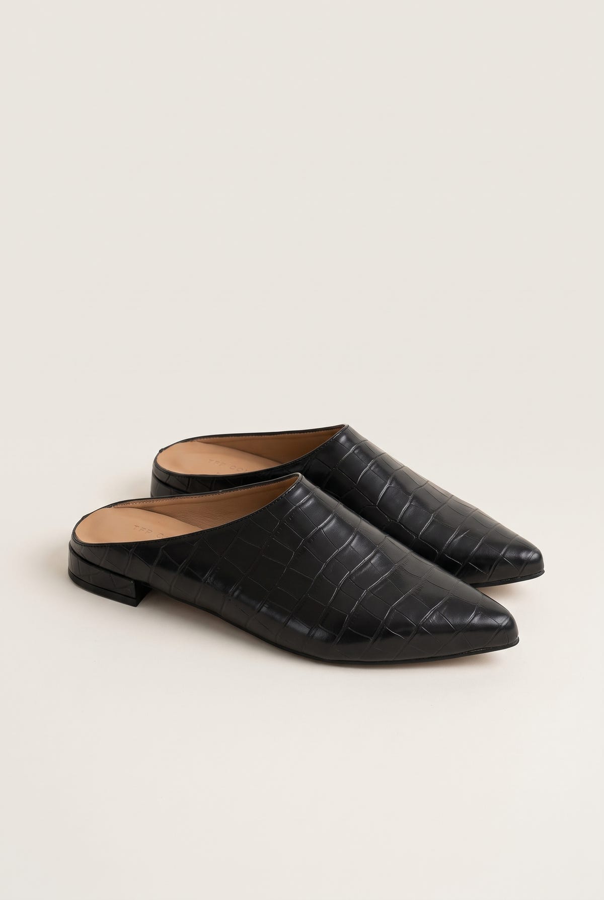Angled view of a pair of black croc-embossed pointed toe mules with open backs and low flat heels
