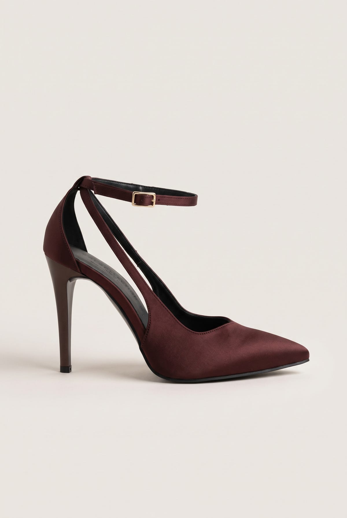 Side view of a brown satin pointed-toe pump with a thin ankle strap and a cut-out side on a pale beige surface