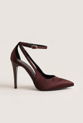 Side view of a brown satin pointed-toe pump with a thin ankle strap and a cut-out side on a pale beige surface