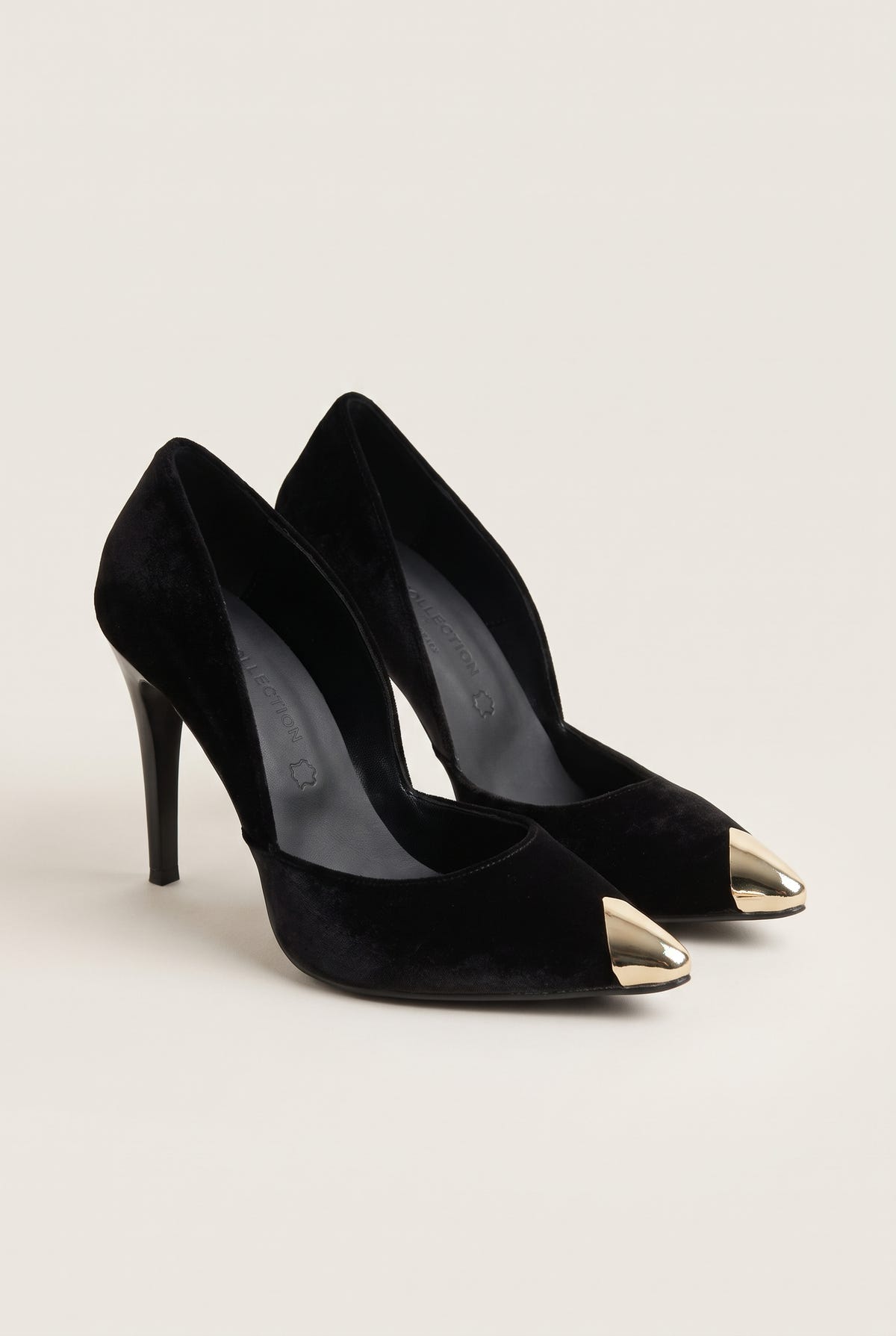 Pair of black velvet pumps seen at an angle with gold metal toe caps and cut-away sides on pale beige