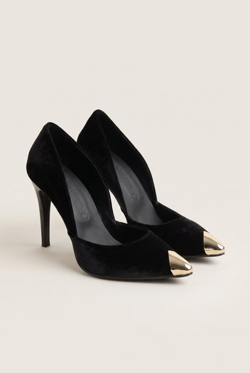 Pair of black velvet pumps seen at an angle with gold metal toe caps and cut-away sides on pale beige