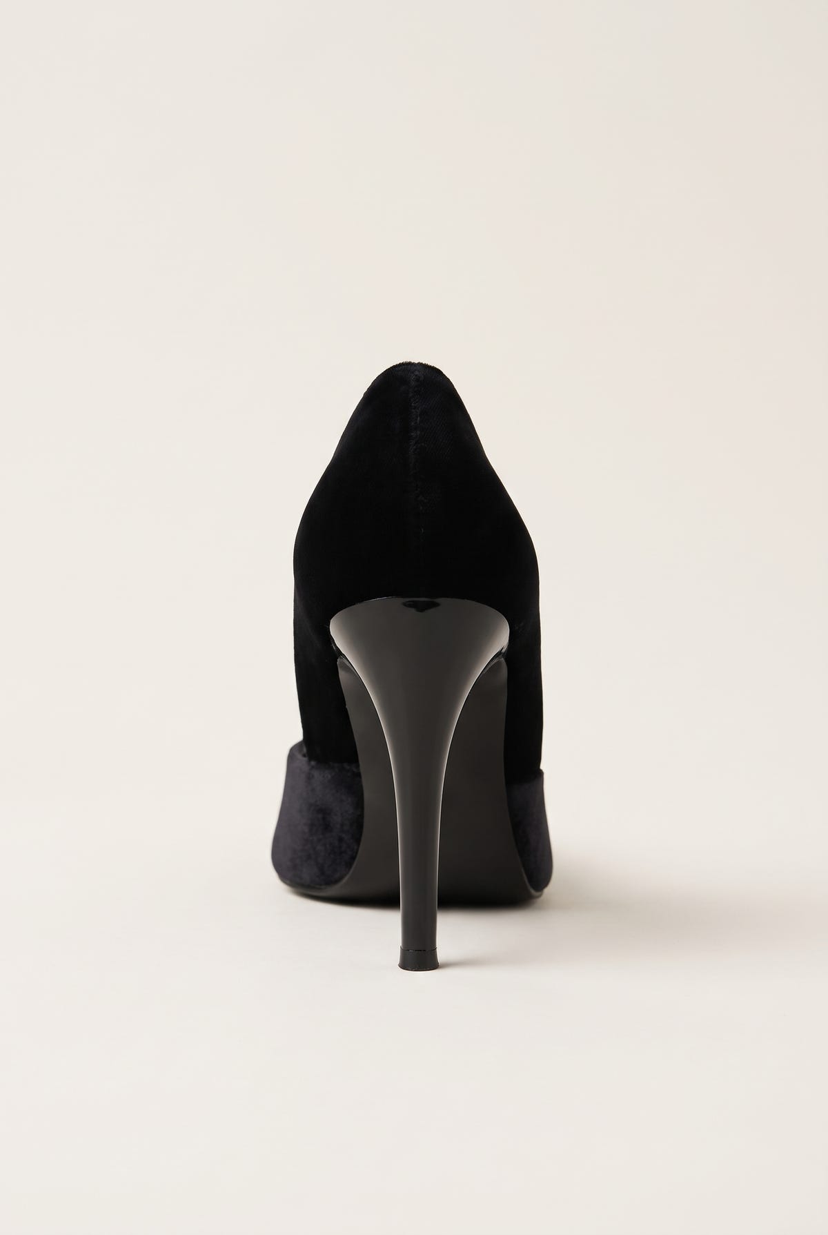 Back view of a black velvet pump with a tall slim heel and smooth rounded heel counter on a pale beige surface
