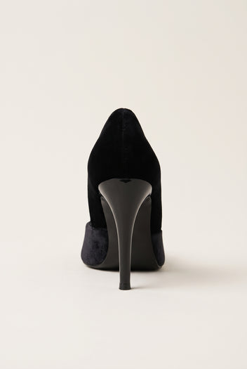 Back view of a black velvet pump with a tall slim heel and smooth rounded heel counter on a pale beige surface