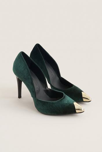 Pair of dark green velvet pumps seen at an angle with gold metal toe caps on a pale beige surface