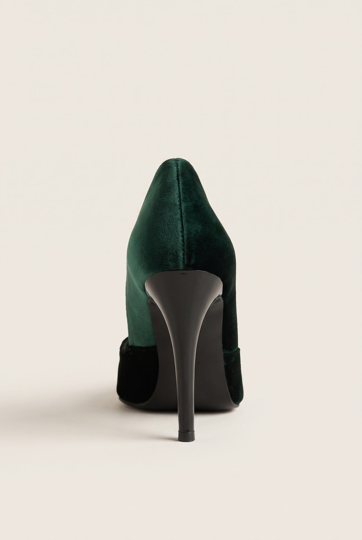 Back view of a dark green velvet pump with a tall slim heel and black heel counter on pale beige