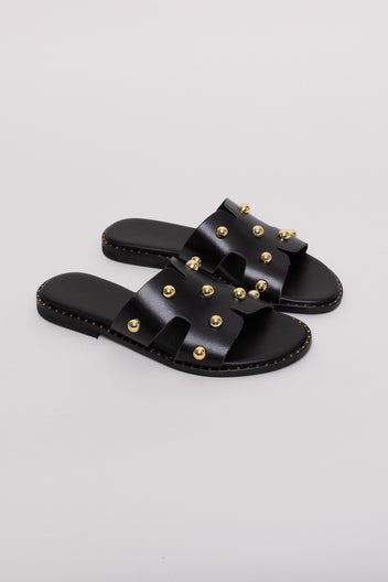 Black leather studded slide sandals with gold studs, angled view on a terracotta backdrop
