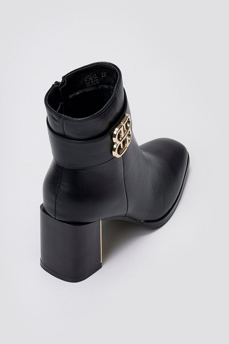 Black leather ankle boot with square toe, block heel and gold-tone logo buckle, angled view on a light grey background