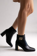 Black leather ankle boots with block heel and gold-tone logo buckle, pair in side view on a model's legs in studio light