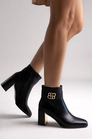 Black leather ankle boots with block heel and gold-tone logo buckle, pair in side view on a model's legs in studio light